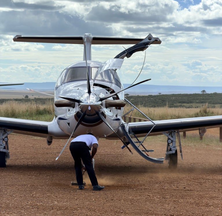 Person standing in front of a white twin-engine aircraft on a dirt runway with grassland and sky in background