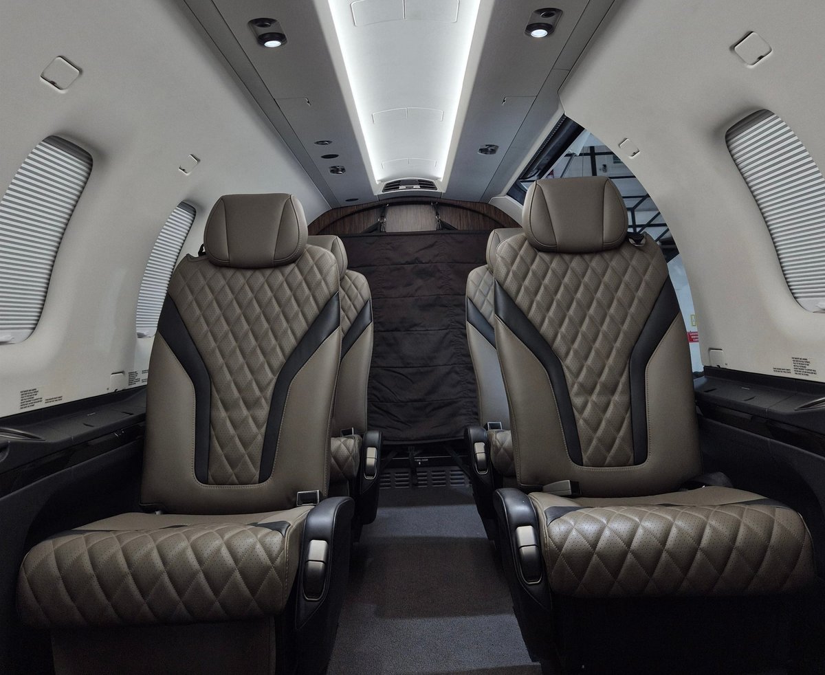 Interior of private jet cabin with quilted leather seats, cabin lighting, and windowed fuselage viewed from rear toward cockpit