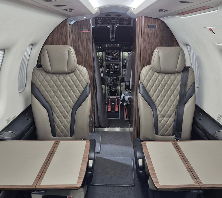 Interior view of a private jet cabin with two beige quilted leather seats, wooden accents, and cockpit visible in the distance