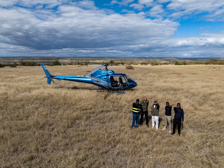 Blue helicopter landed in a dry grassland with a group of people standing nearby under a cloudy sky