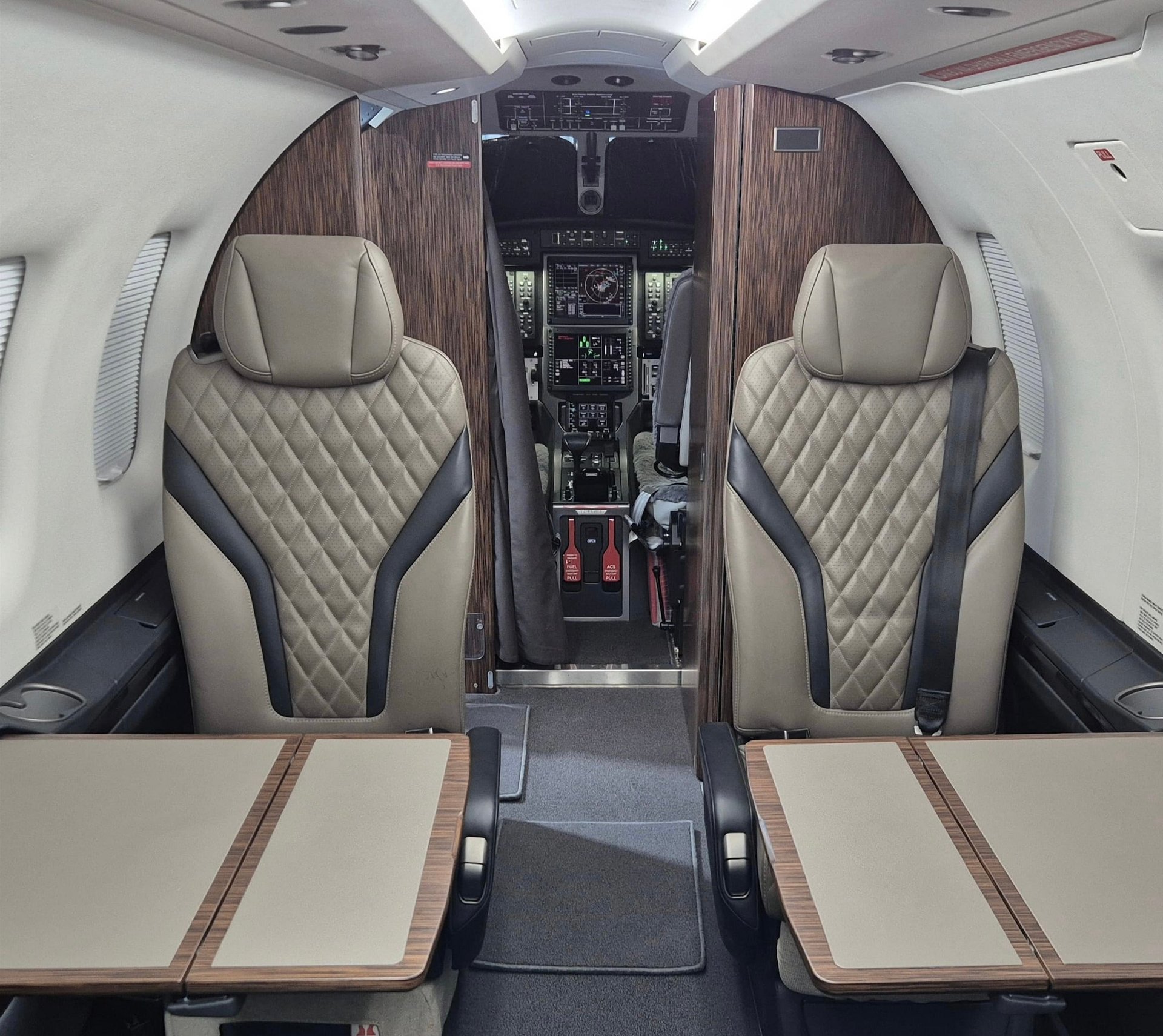 Interior of a private jet cabin with two cream-colored leather seats, dark wood accents, and cockpit visible in background