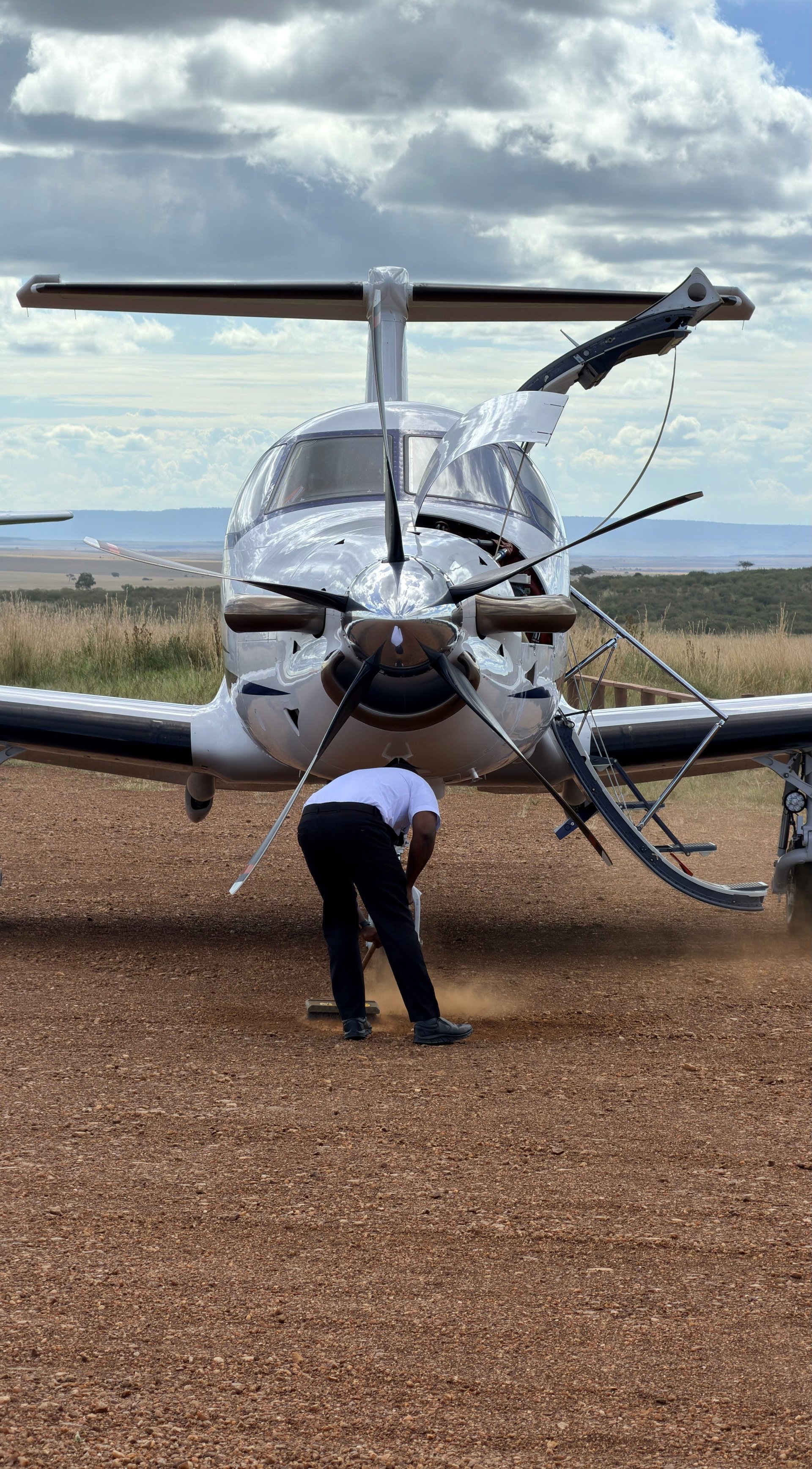 A person in formal attire stands under the tail rotor of a white twin-engine helicopter parked on red dirt airstrip with cloudy sky and savanna landscape in background