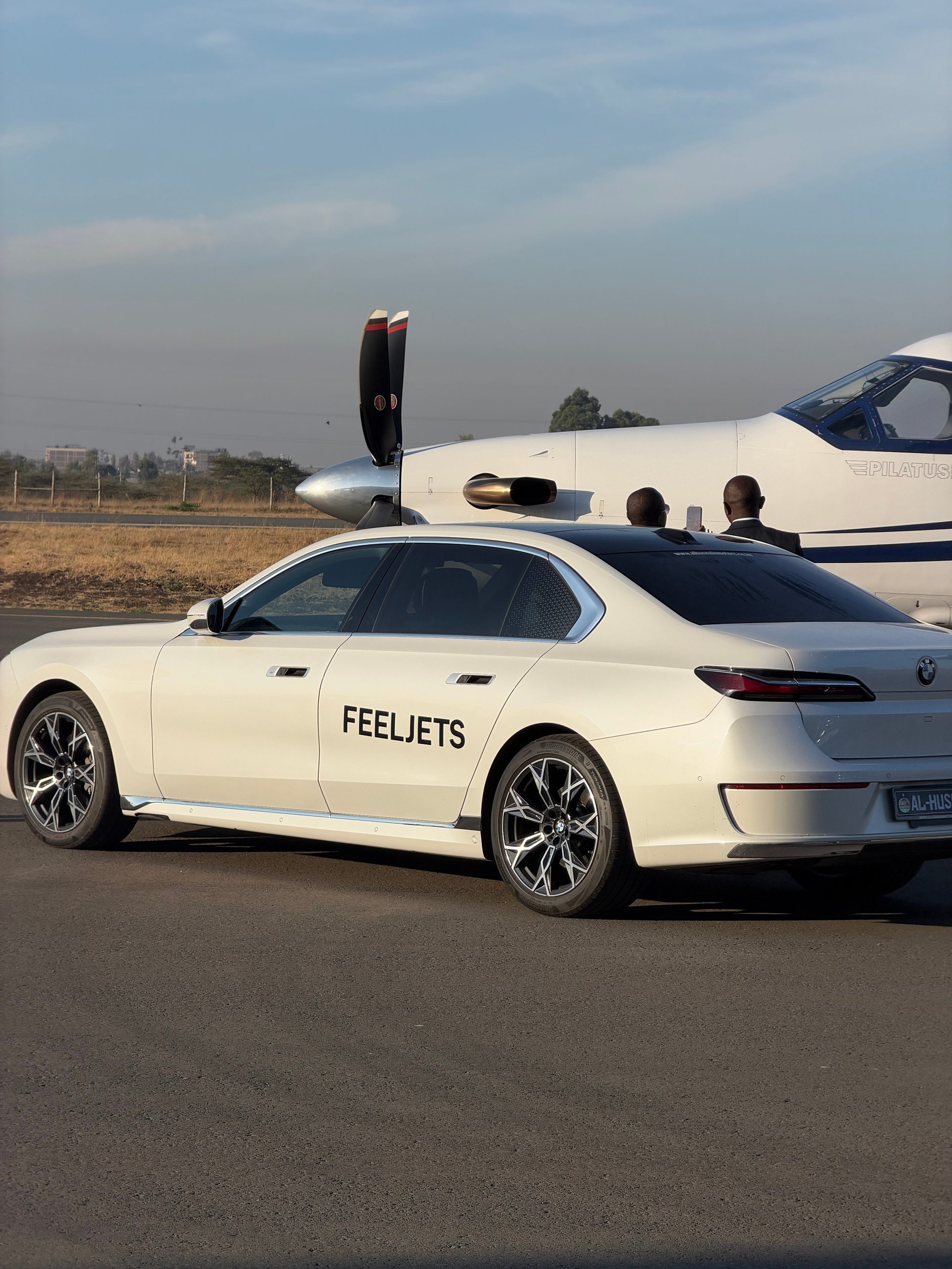 White BMW sedan with Feeljets branding parked in front of a small private aircraft on a tarmac