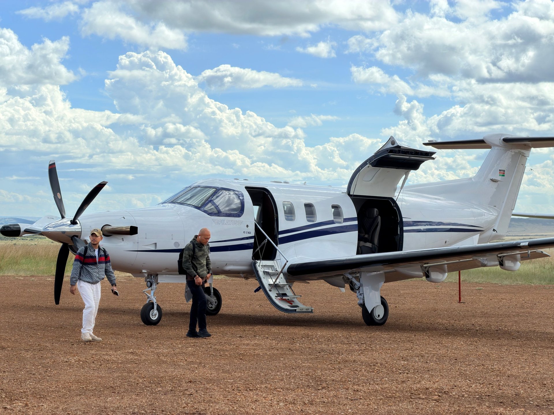 A white twin-engine turboprop aircraft parked on a dirt runway with three people standing nearby and cloudy sky in the background