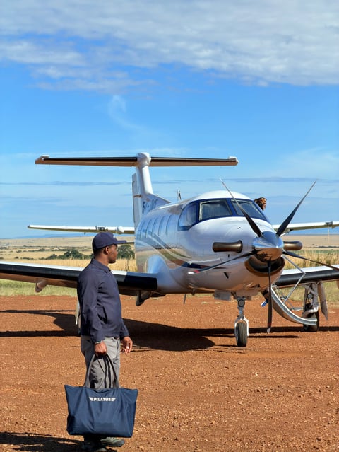 Man in black jacket carrying blue tote bag stands in front of white and blue twin-engine aircraft on red dirt runway under clear blue sky