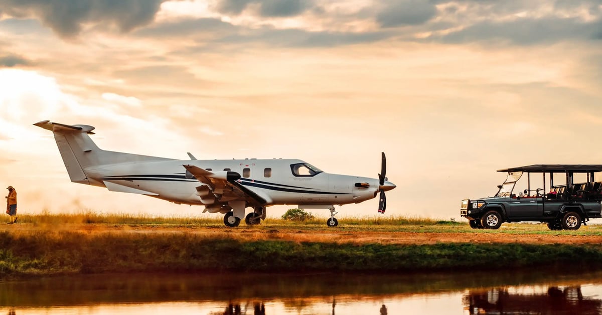 Single-engine propeller airplane parked on dirt runway with safari vehicle and people nearby at sunset